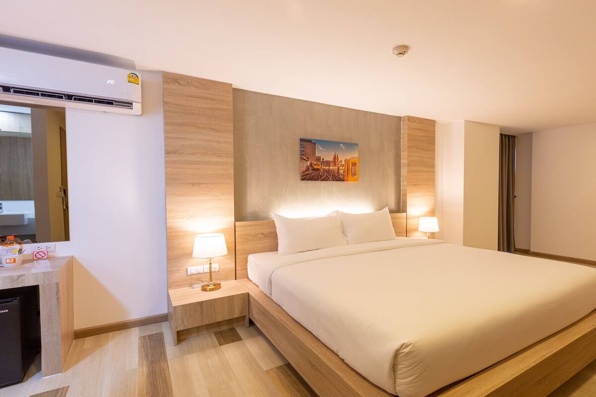 A modern hotel room with a large bed, bedside lamps, a painting on the wall, and an air conditioning unit.