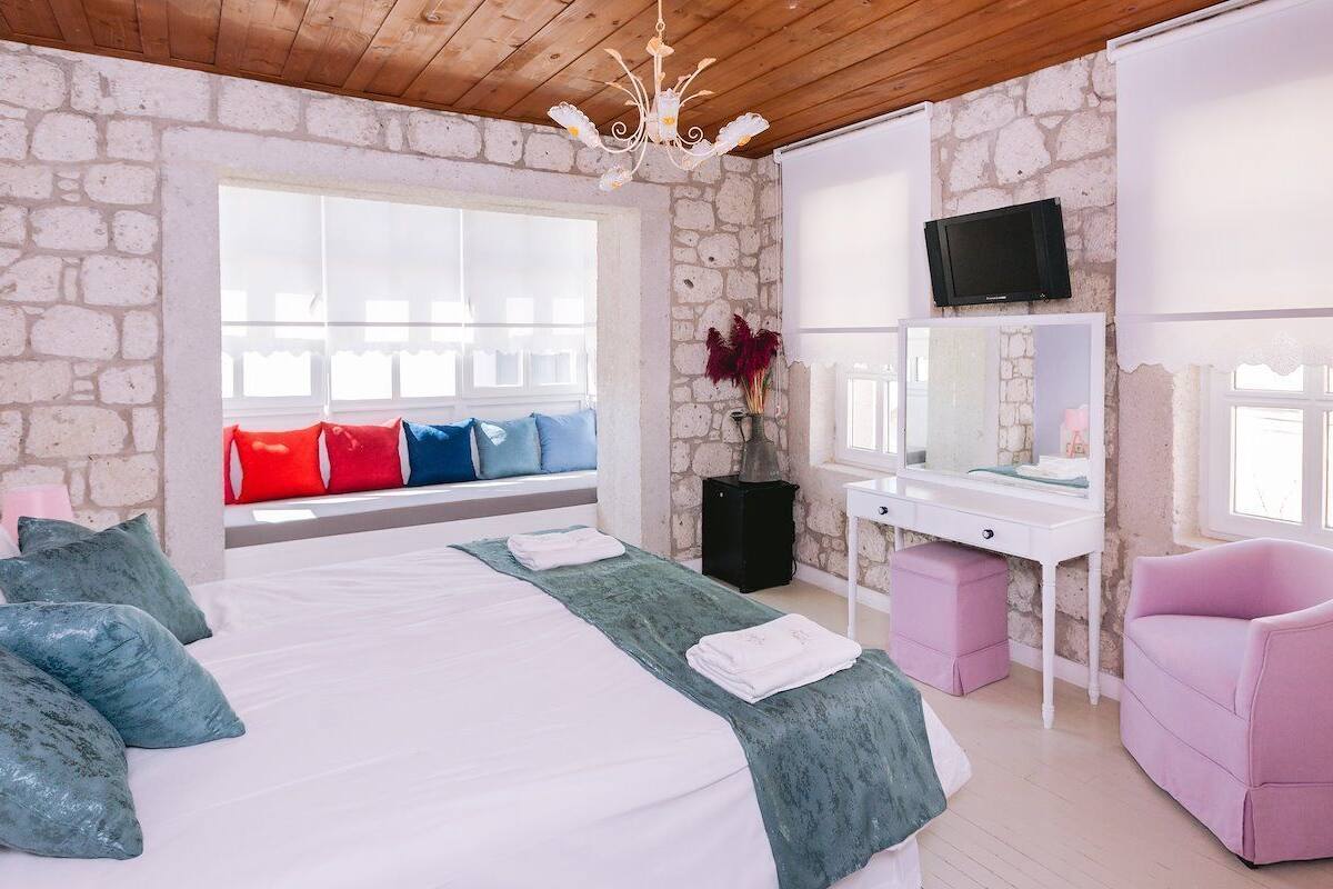 Un dormitorio con pared de piedra, cama con muchos almohadones, escritorio con espejo, una silla rosa y televisor en la pared.
