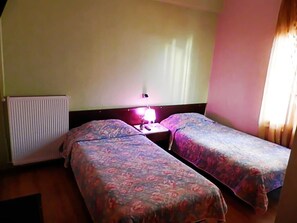 Una habitación con dos camas individuales, una mesita de noche con lámpara, un radiador y una ventana con cortinas.