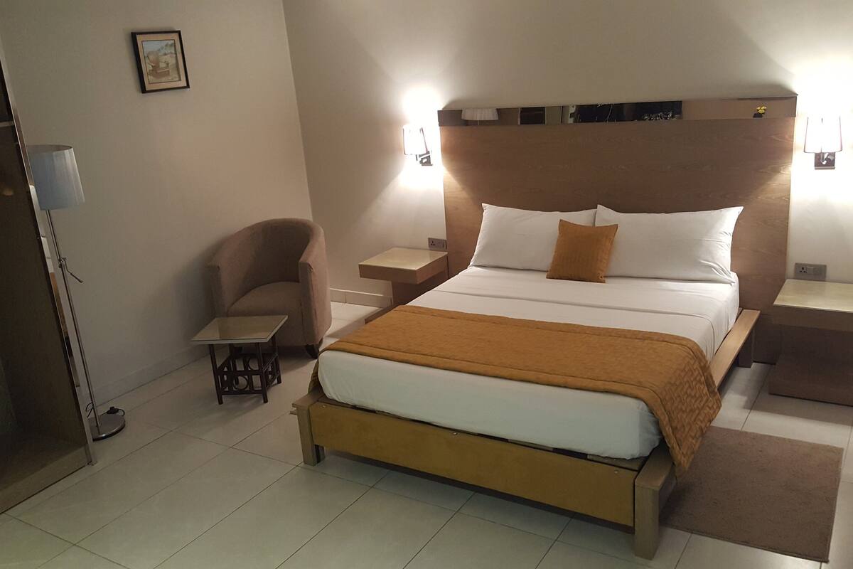 Una habitación de hotel ordenada con una cama grande, una silla, una mesita pequeña y lámparas de noche.