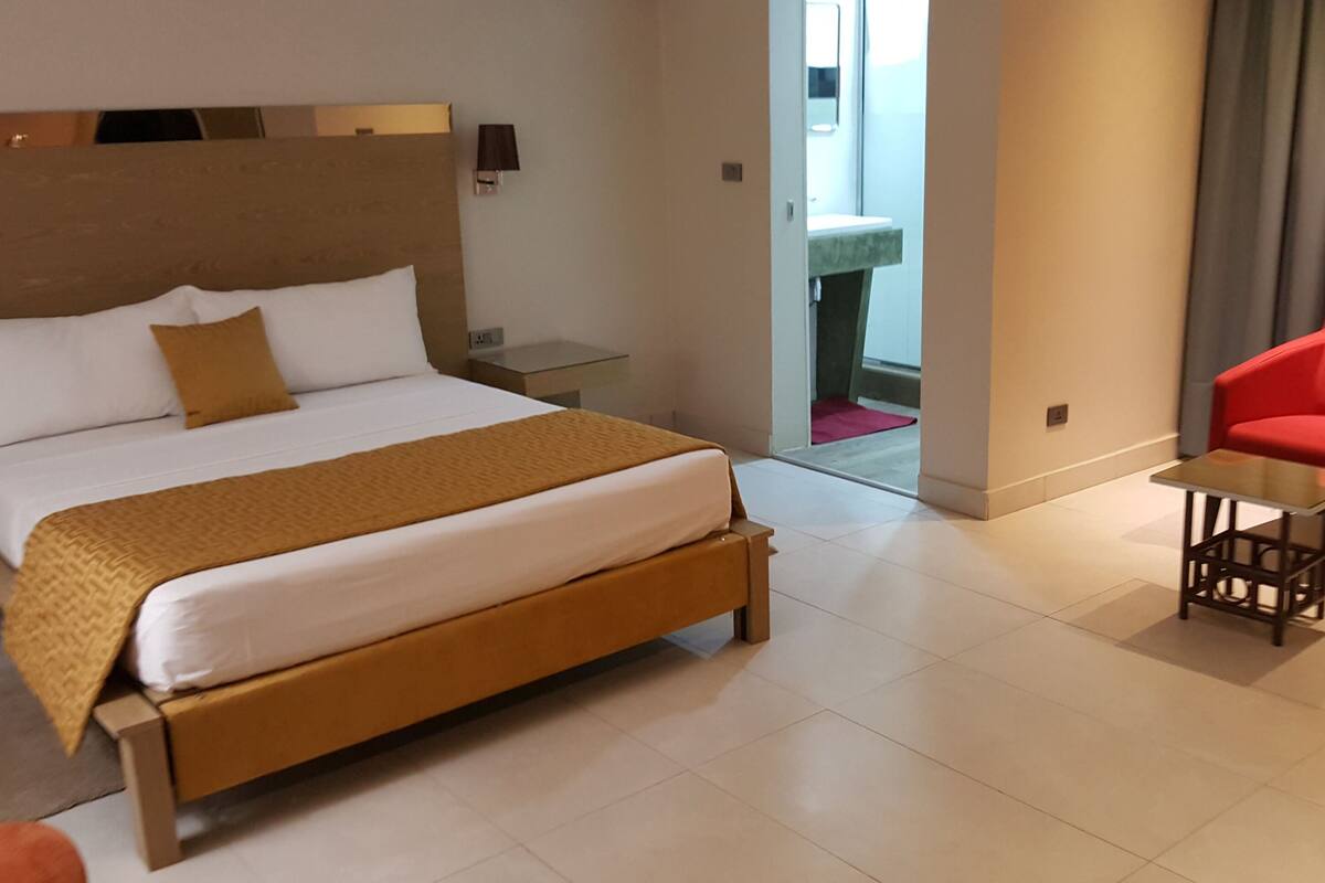Habitación de hotel moderna con una cama grande, una mesita pequeña, una silla roja y un baño visible a través de una puerta abierta.