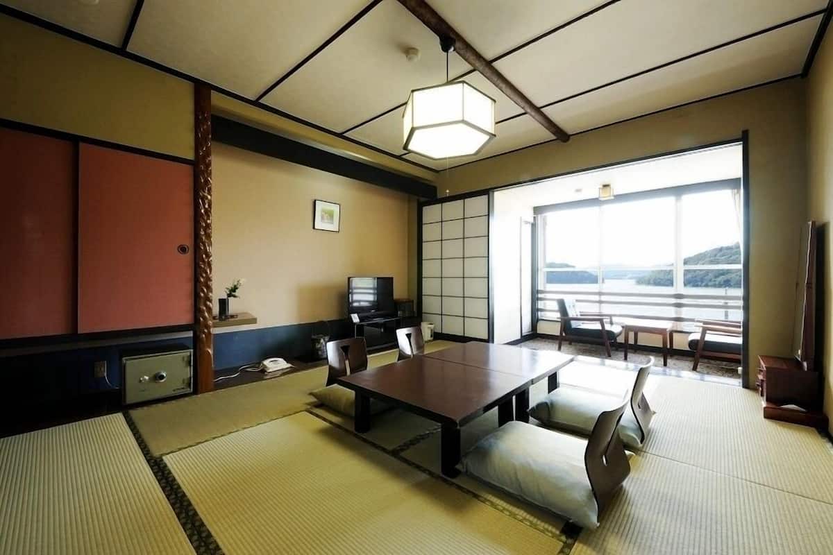 A traditional Japanese room with tatami flooring, a low table, chairs, a television, and a view of a mountainous landscape through large windows.
