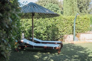 Un jardín con dos tumbonas bajo un paraguas grande, rodeado de follaje.