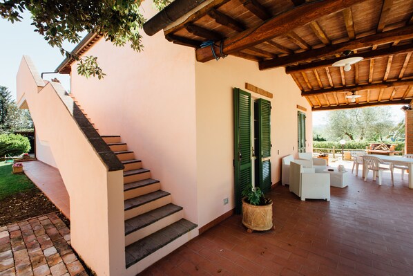 Un patio exterior techado con techo de madera, una escalera que conduce a una puerta con contraventanas verdes y un área de descanso con mobiliario blanco.