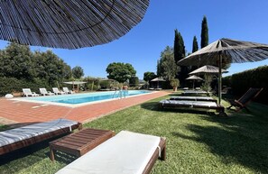 Piscina con sillones de descanso y sombrillas, rodeada de césped y árboles.