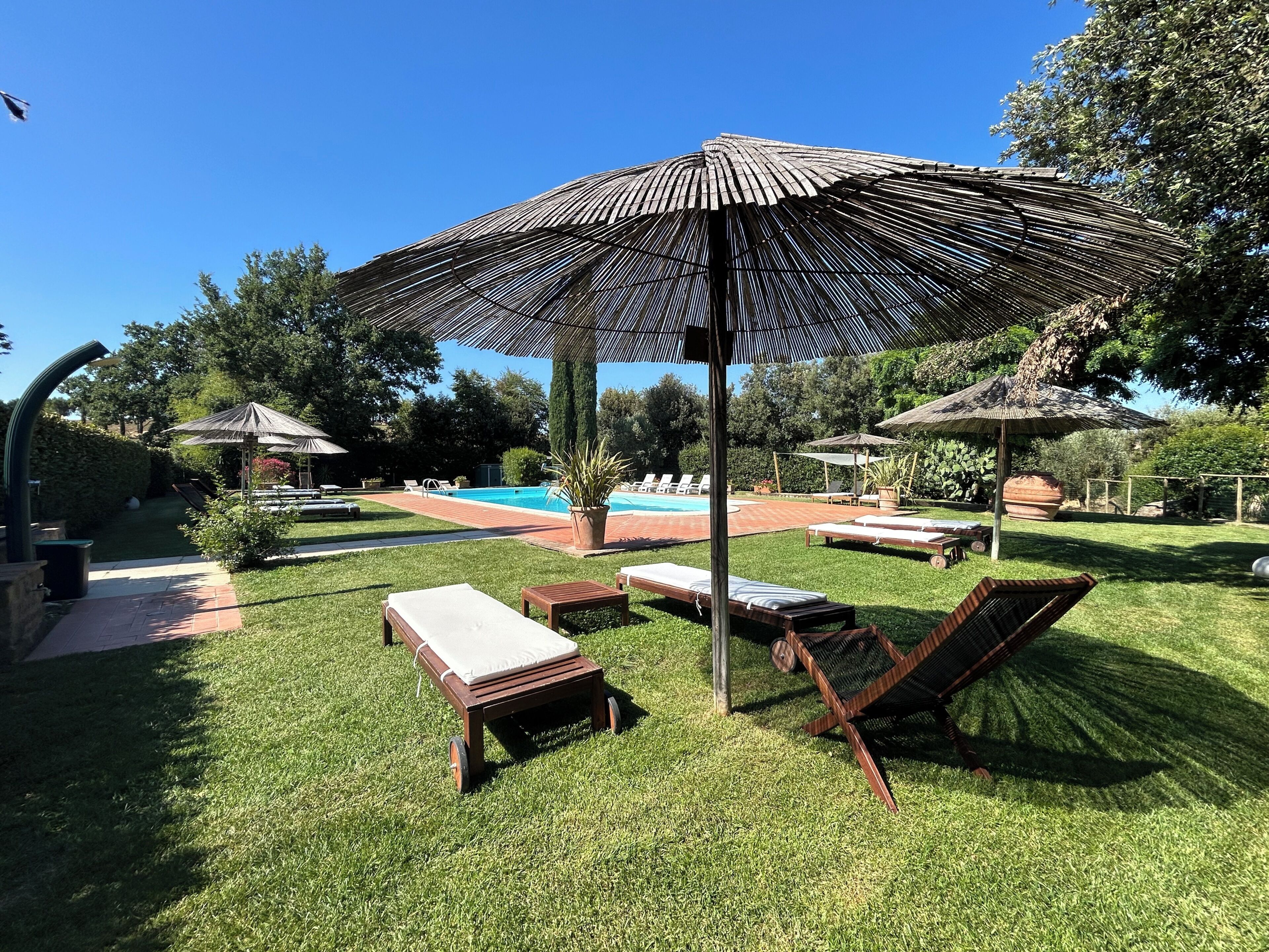 Zona junto a la piscina con sillones de descanso, una terraza de madera y una sombrilla grande.