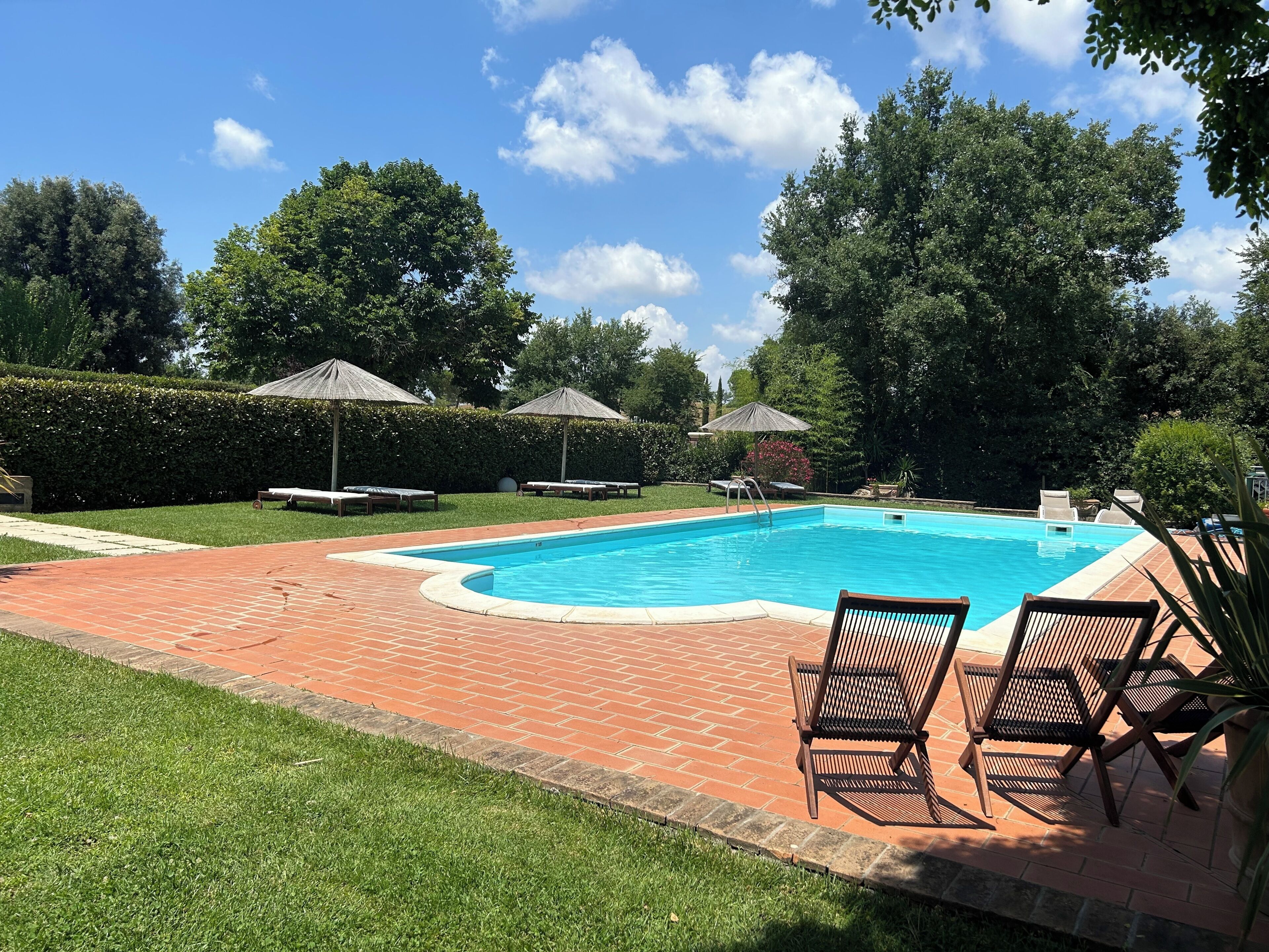 A rectangular swimming pool with a brick border, surrounded by grass and trees.