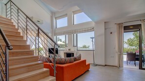 A spacious living area with a wooden staircase, a bright orange sofa, and large windows offering natural light and views. - Villa retreat in the heart of Cretan nature (Rethimno)