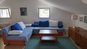 A modern living room with a sectional sofa, a wooden coffee table, and a small cabinet. There are two windows providing natural light, and a painting is visible on the wall. - Apartment Luna (Konavle)