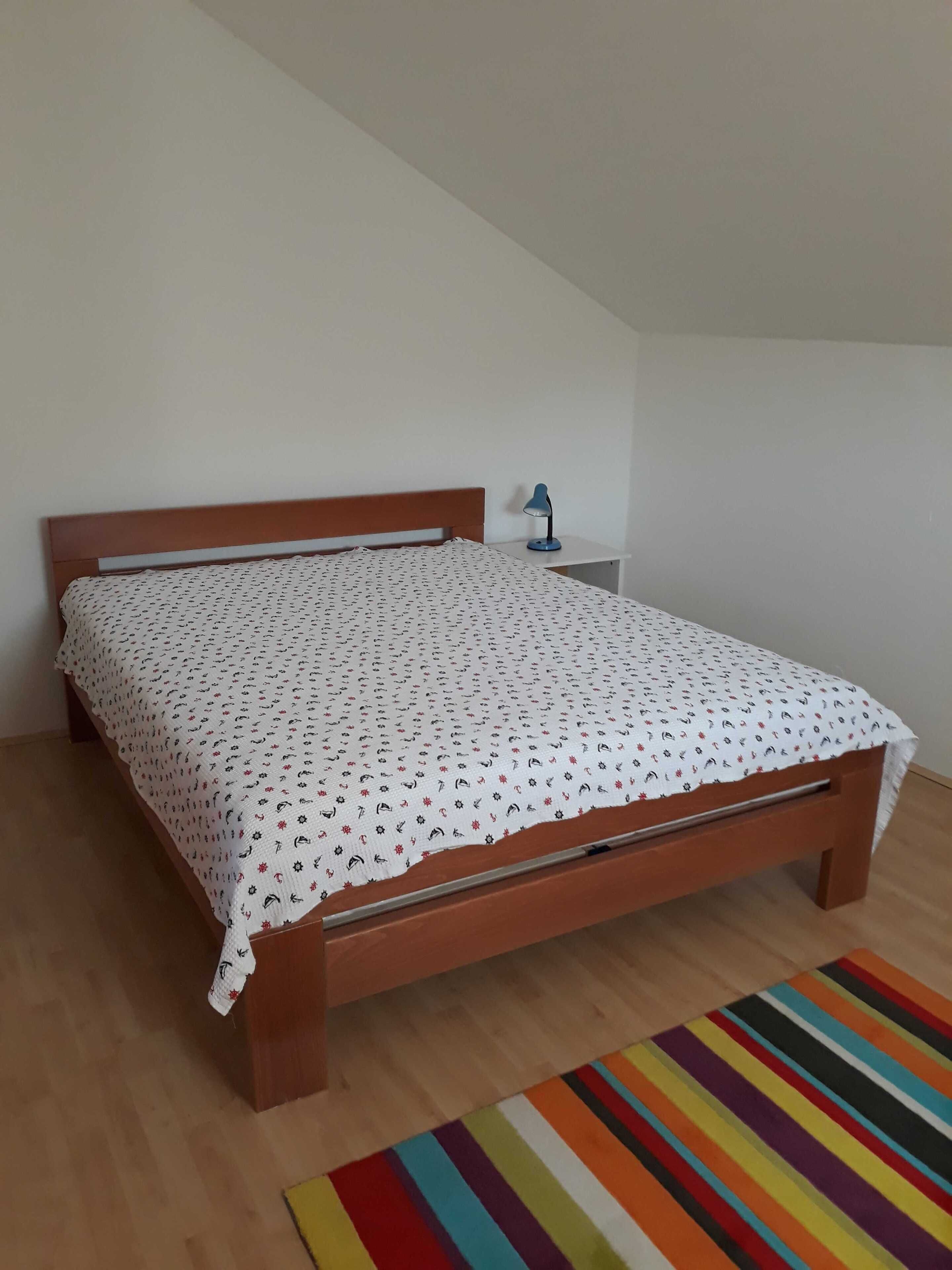 Ein Einzelbett mit gemustertem Überwurf, ein Nachttischchen mit einer Lampe und ein farbenfroher Streifen Teppich auf Holzdielen in einem Zimmer mit weißen Wänden.