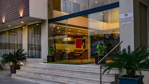 Entrada de hotel com fachada de vidro, escadaria que leva à entrada e plantas em vasos de ambos os lados.