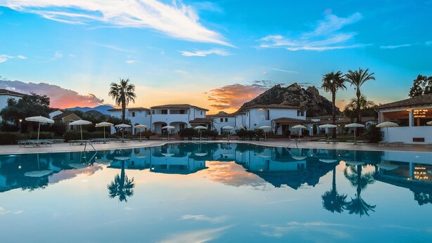 Una piscina con sillas de descanso y sombrillas, rodeada de edificios blancos y palmeras, que refleja el cielo al atardecer.