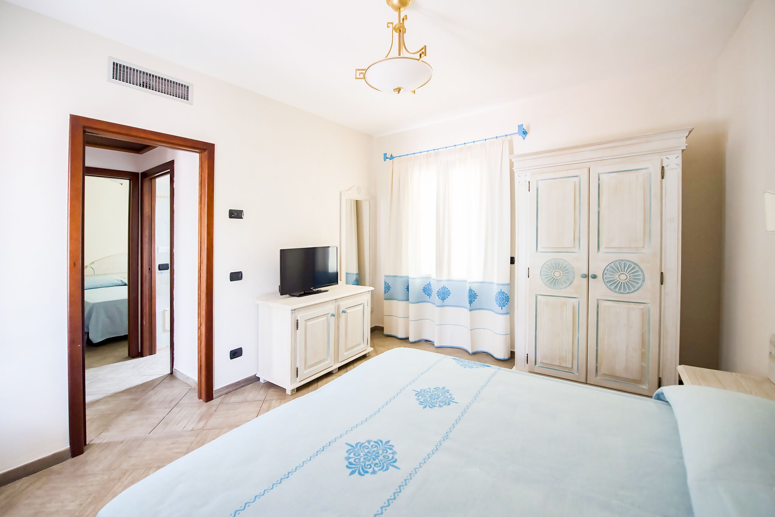A bedroom with a bed, a TV on a stand, a wardrobe, and a window with curtains.