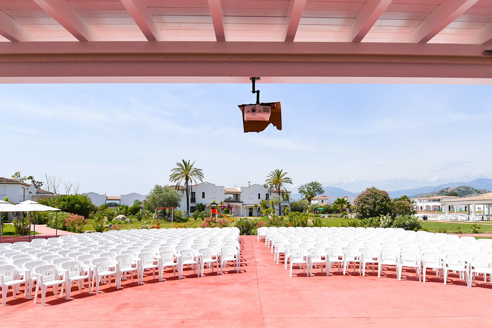 An outdoor seating area with rows of white plastic chairs and a projector mounted on the ceiling.