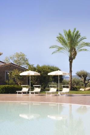 A pool area with lounge chairs and umbrellas, a palm tree, and a stone building in the background. - Marina Torre Navarrese Resort (Lotzorai)