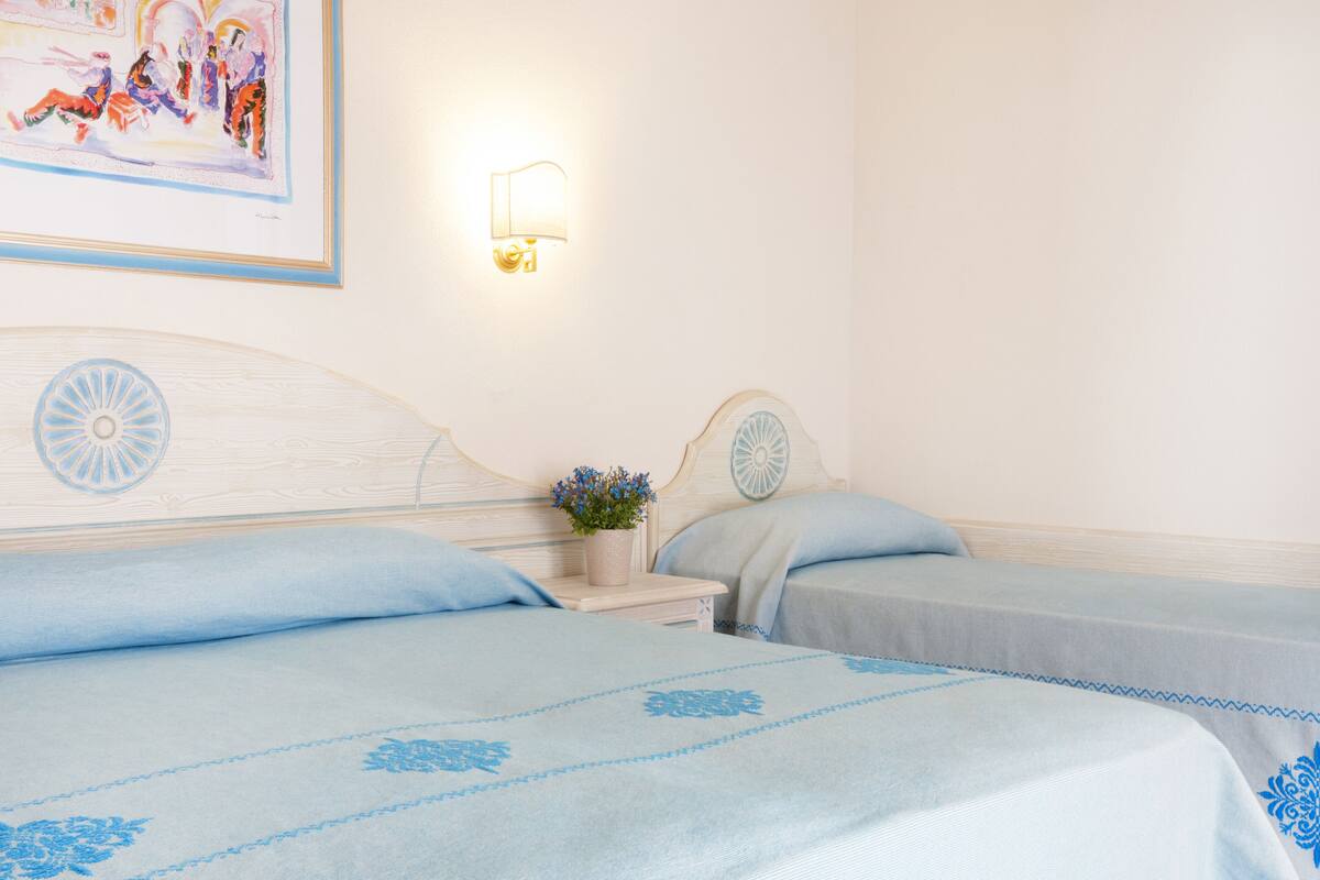 Ein Hotelzimmer mit zwei Betten, jedes mit einer blauen, blumigen Tagesdecke, einem kleinen Nachttisch mit einer Pflanze und einer wandmontierten Lampe.