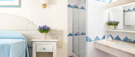 A hotel room with a bed, bedside table, and a small cabinet. There is a shower area with a glass door and a sink with a mirror above it.