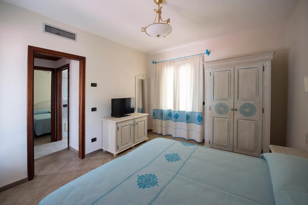 Une chambre à coucher avec un lit, un téléviseur posé sur un meuble, une armoire et une porte donnant sur une autre pièce.