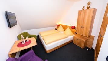 Une petite chambre à coucher, bien rangée, avec un lit, une armoire en bois, une petite table surmontée d’un set de table rouge et une chaise violette.