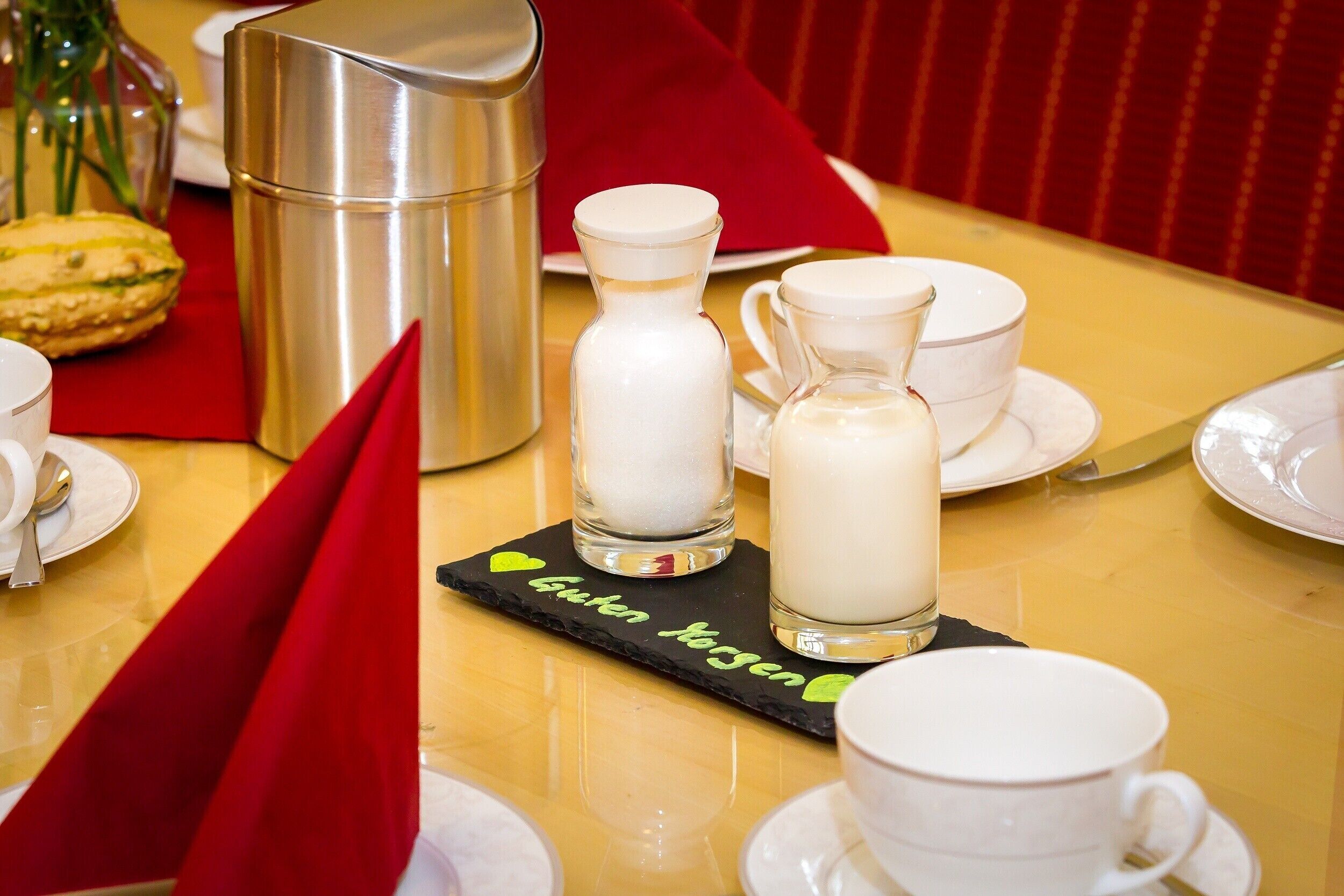 Une table dressée avec des tasses et des soucoupes blanches, un thermos en argent et deux bouteilles en verre, le tout posé sur un dessous de verre noir orné de texte.