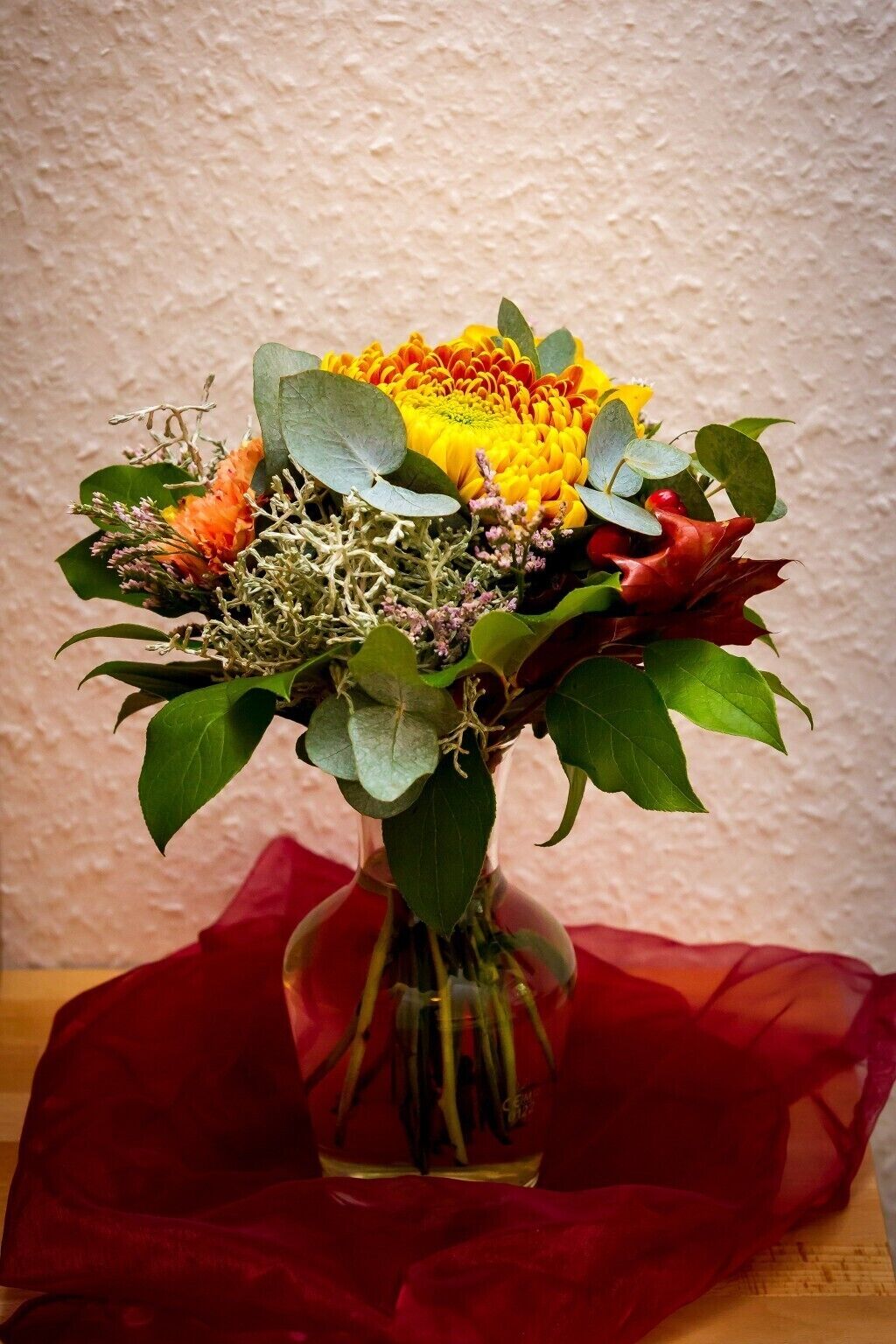 A bouquet of flowers in a clear glass vase on a wooden surface with a red cloth underneath.