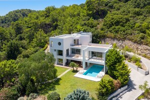 Uma casa moderna de dois andares com piscina, rodeada por vegetação exuberante e uma paisagem montanhosa.
