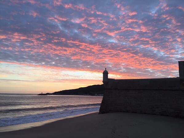 Un paysage côtier avec un mur de pierre et la silhouette d’un phare se découpant sur un ciel au coucher du soleil.
