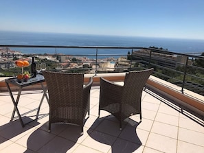Un balcon avec des fauteuils en osier et une table où sont disposés des fruits et une bouteille, offrant une vue sur le paysage urbain de la côte.