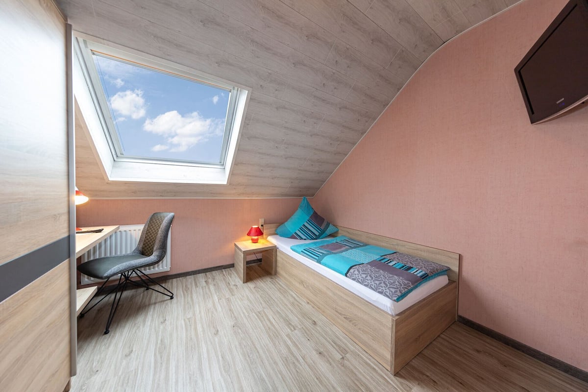 Una habitación pequeña y acogedora con techo inclinado, tragaluz, cama con almohadas, escritorio con silla y televisor instalado en la pared.
