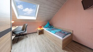 A small, cozy room with a sloped ceiling, a skylight, a bed with a blue and gray patterned bedspread, a desk with a chair, and a small bedside table with a lamp.