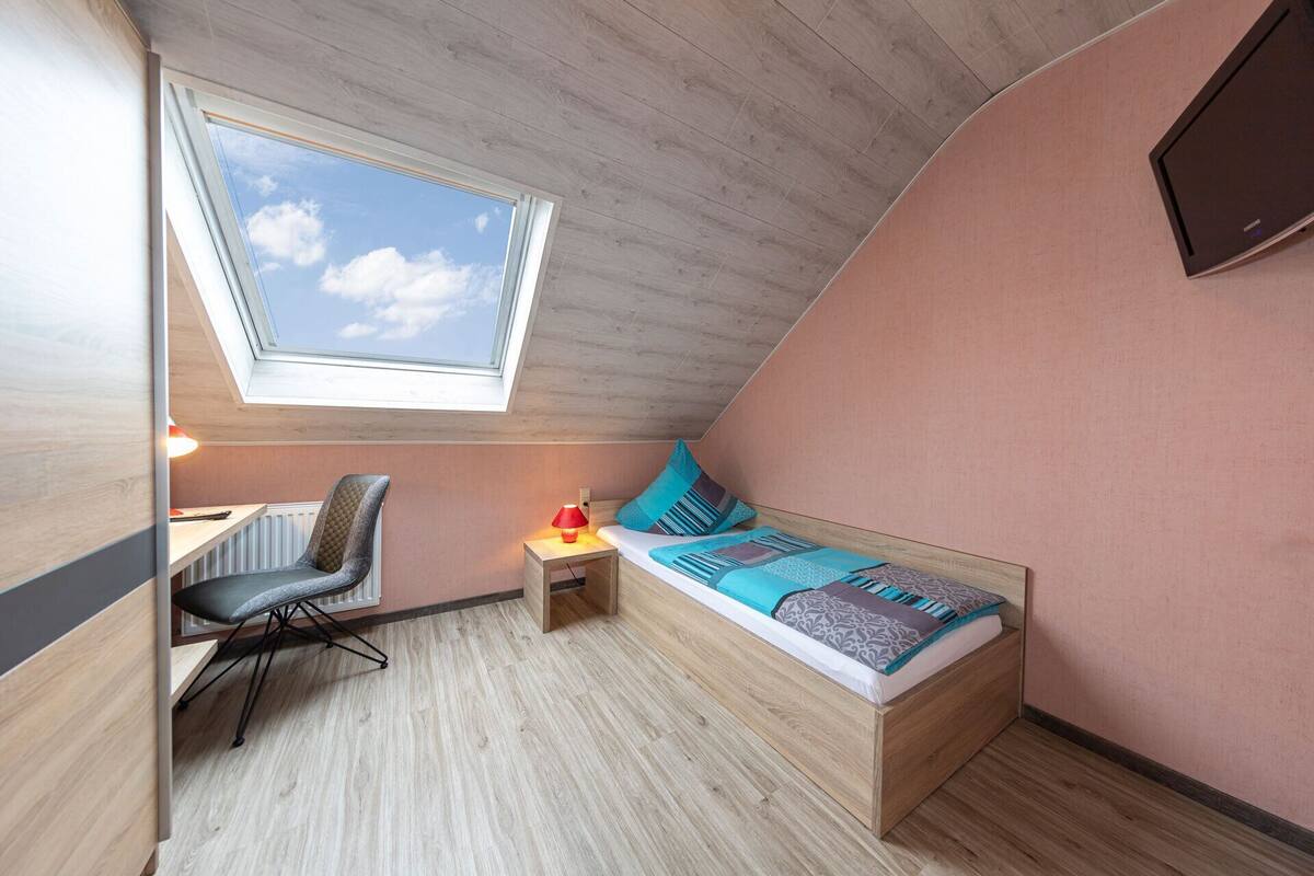 Una habitación pequeña y acogedora con techo inclinado, tragaluz, cama con almohadas, escritorio con silla y televisor instalado en la pared.