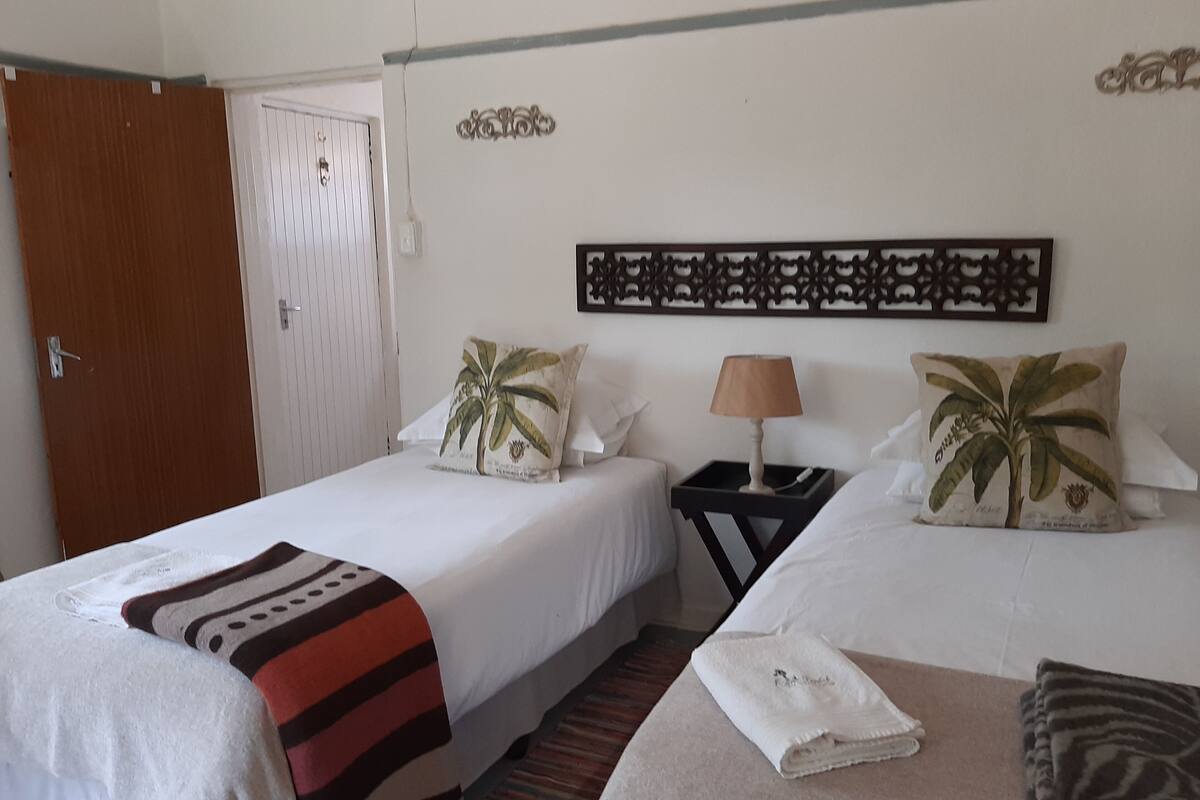 A hotel room with two single beds, a wooden door, a decorative wall piece, and a nightstand with a lamp.