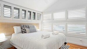 Une chambre à coucher avec un lit, des stores blancs, une table de chevet et une lampe.