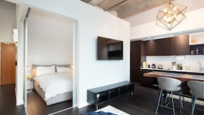A modern hotel room with a bed, a TV mounted on the wall, a dining area with a bench and chairs, and a kitchen area with cabinets and a countertop. - Hudson Hideaway-Historic Hudson Building in Beautiful Downtown Victoria by Victoria Prime! (Victoria)