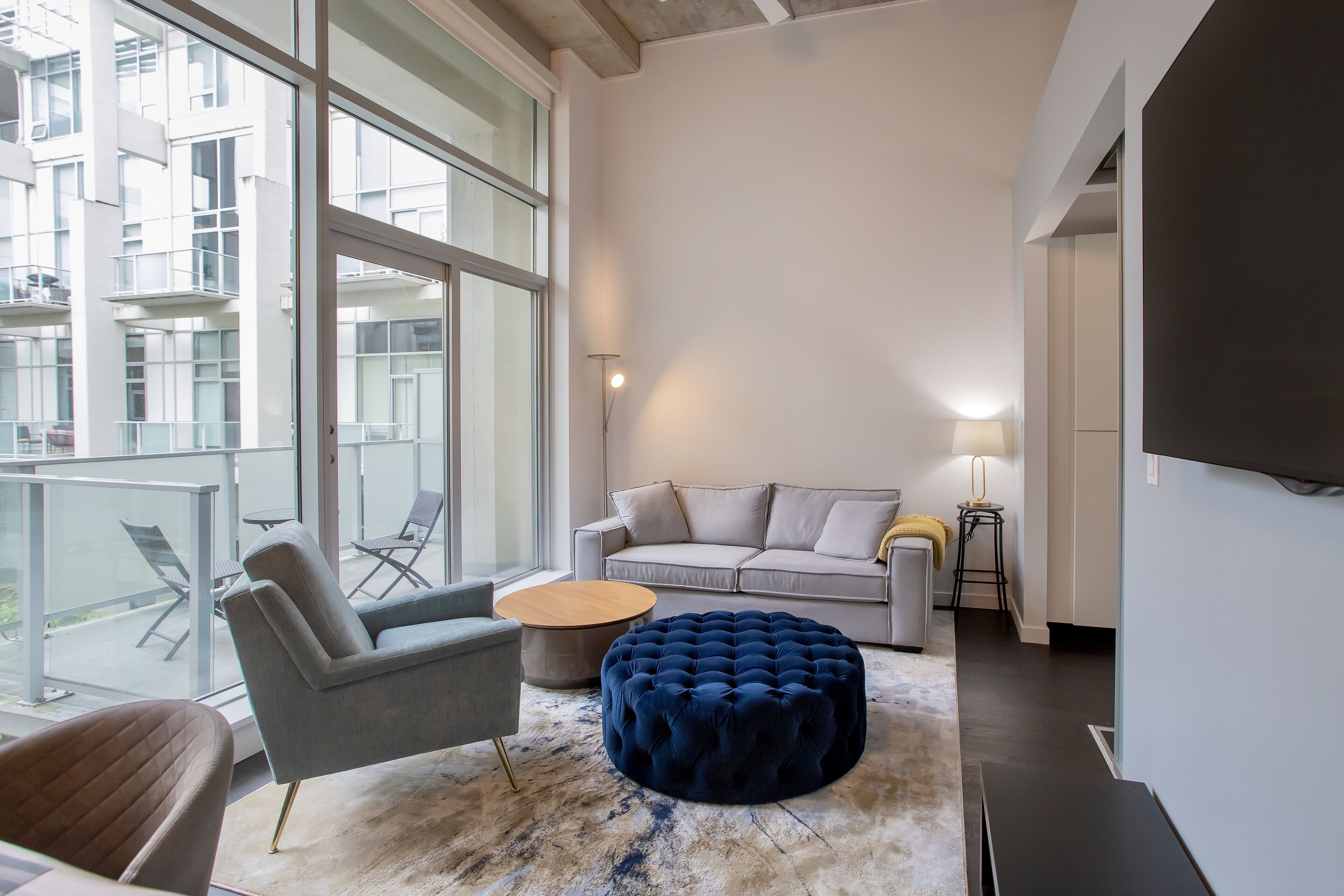Ein modernes Wohnzimmer mit einer Couch, einem Sessel, einem Hocker und einem runden Couchtisch. Große Fenster bieten Blick auf die Stadtlandschaft.