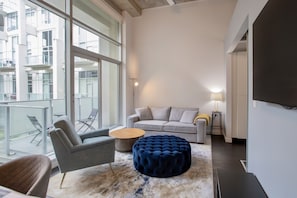 Un salon moderne comprenant un canapé, un fauteuil, un pouf et une table basse ronde. De grandes fenêtres offrent une vue sur le paysage urbain.