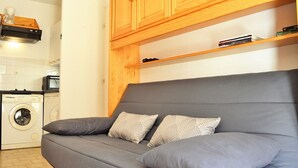 Ein kompakter Wohnbereich mit grauem Sofa, zwei gemusterten Kissen, einer Waschmaschine und Holzschränken.