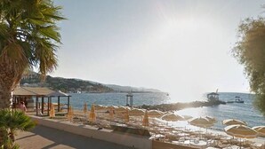 Blick auf einen Strand mit Sonnenschirmen, einer Holzterrasse und Meer sowie Booten.