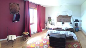 Een slaapkamer met een bed, een stoel, een bureau, een nachtkastje, een lamp en een raam met gordijnen.