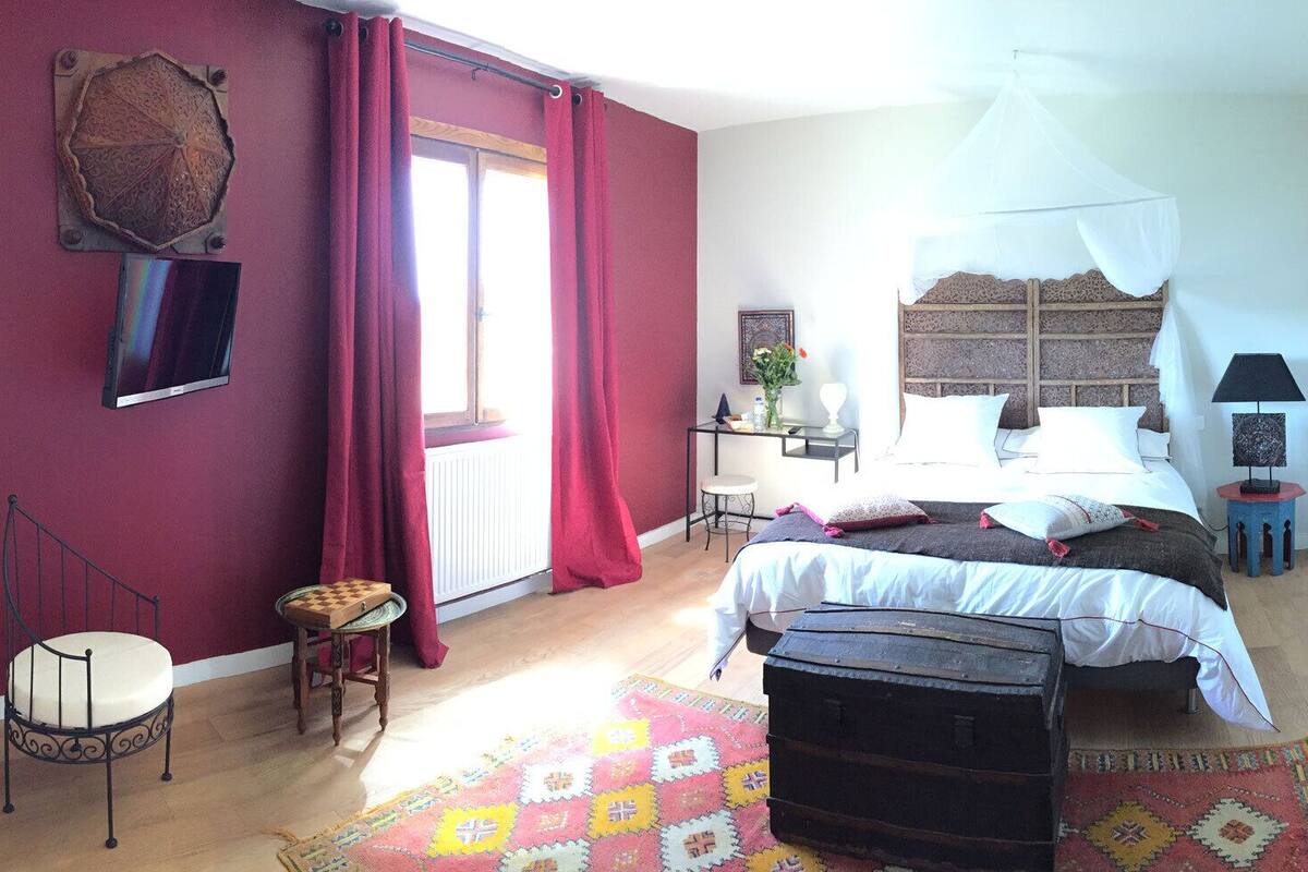A bedroom with a bed, a chair, a desk, a bedside table, a lamp, and a window with curtains.