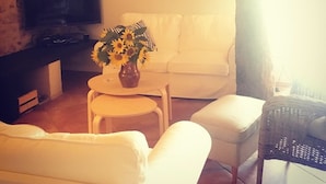 Een woonkamer met een witte bank, een kleine ronde tafel met een vaas zonnebloemen, een televisietoestel op een standaard en een rieten fauteuil.
