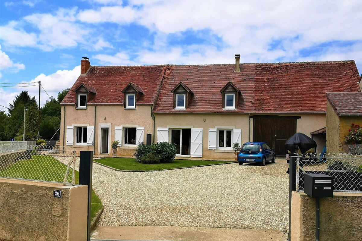 Une maison à deux étages, avec un toit recouvert de tuiles rouges, une allée en gravier et une voiture bleue garée dans l’allée.