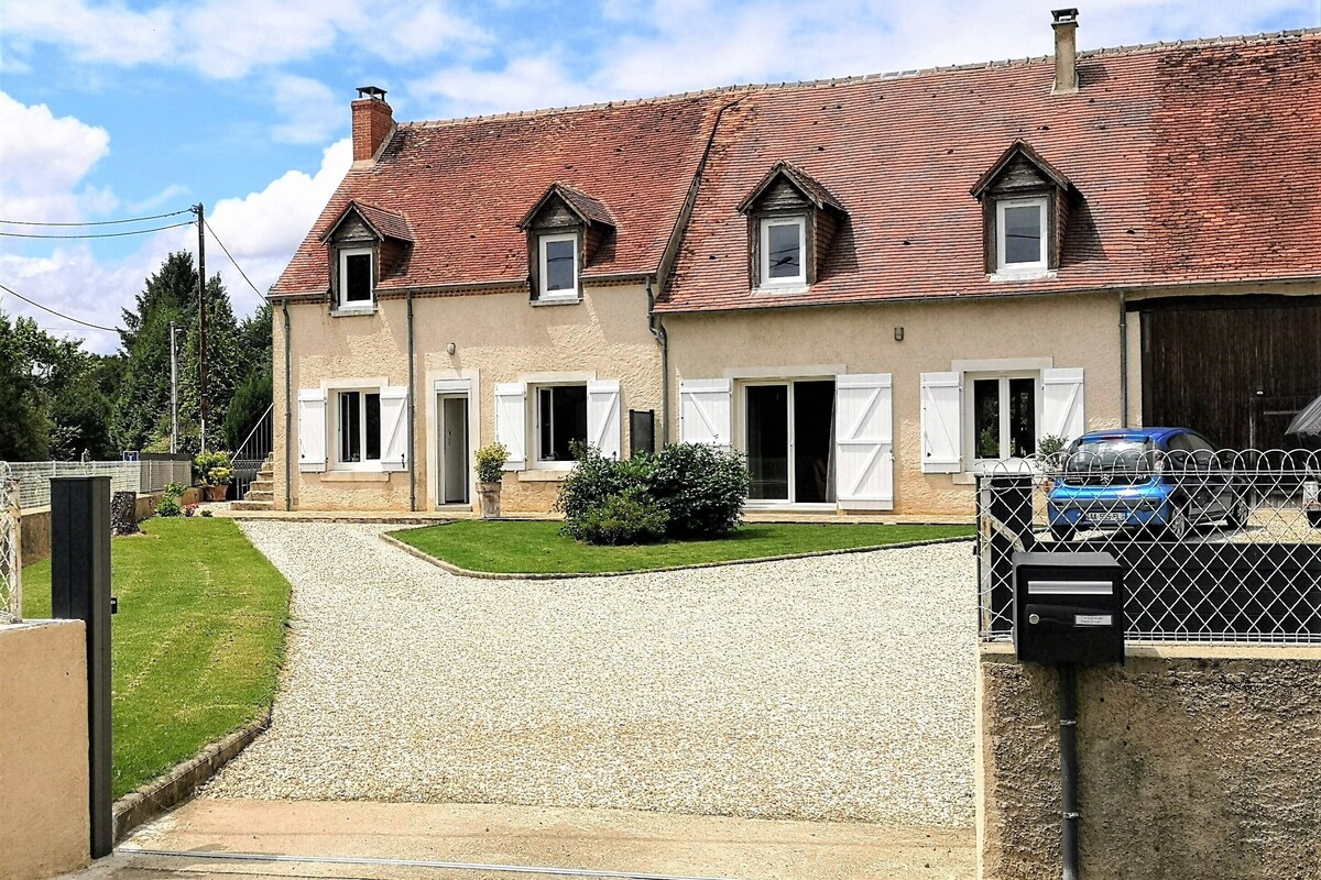 Une maison à deux étages, avec un toit recouvert de tuiles rouges, des volets blancs et une allée gravillonnée.