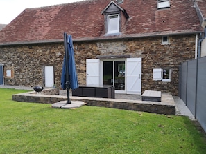 Un bâtiment en pierre avec un toit recouvert de tuiles rouges, un patio couvert aménagé avec du mobilier d’extérieur et une cour gazonnée.