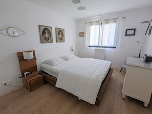 Une chambre à coucher avec un lit en bois, du linge de lit blanc, une table de chevet avec une lampe, une commode et deux tableaux encadrés accrochés au mur.