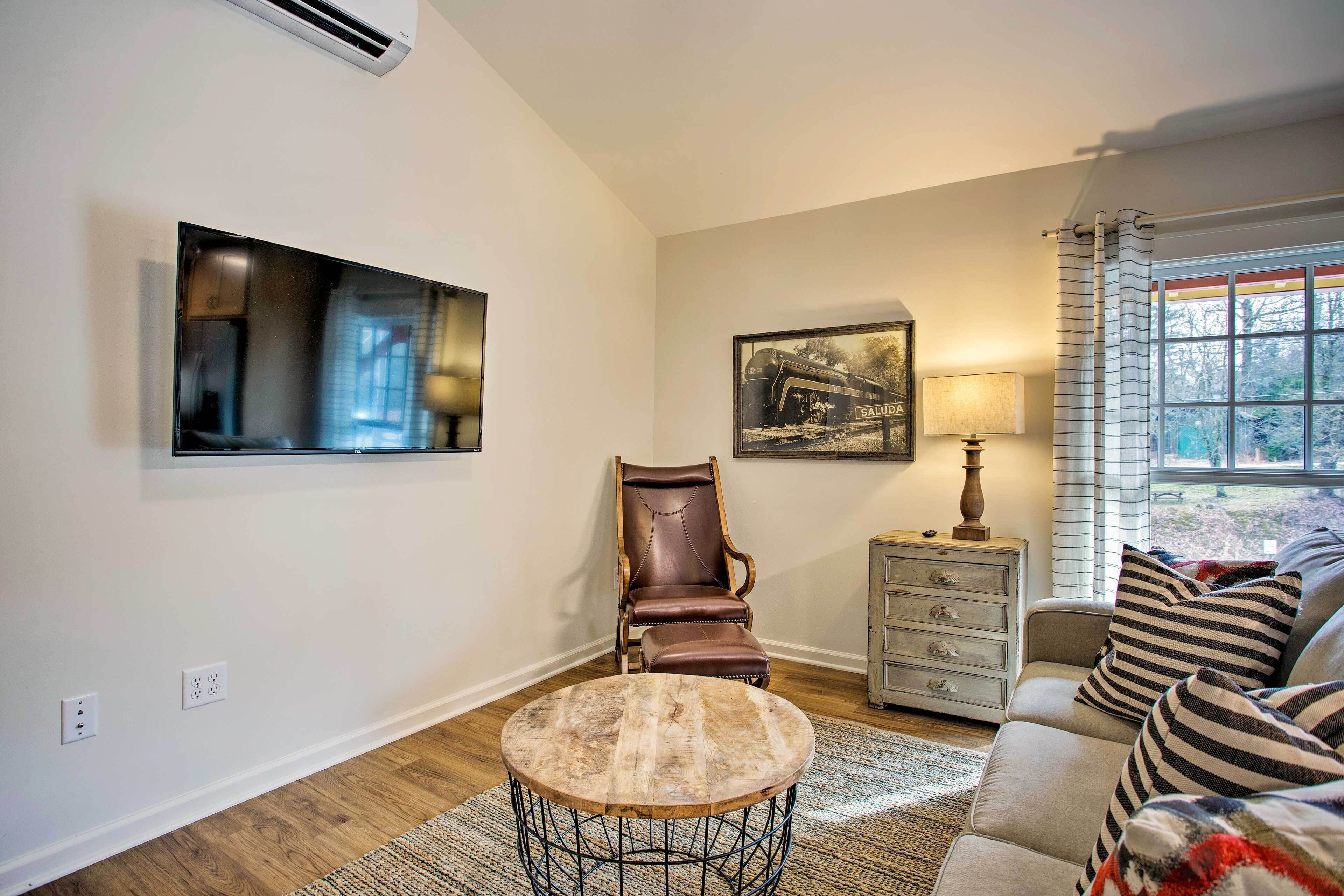 A living room with a round coffee table, a brown leather chair, a sofa with striped pillows, a framed picture on the wall, and a window with curtains.