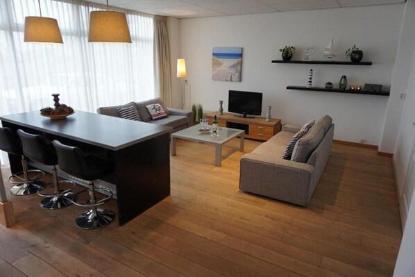 A modern living room with a wooden floor, a grey sofa, a coffee table, a TV, and a bar area with stools.