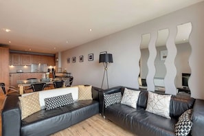 A modern living room with a black leather sofa, a dining area, and a kitchen in the background.