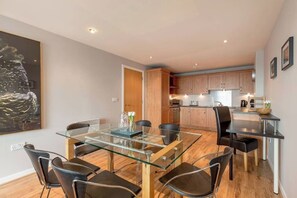 A modern dining area with a glass table and chairs, a kitchen with wooden cabinets, and a painting on the wall. - Close to Hydro/SECC: Modern Riverside Apt with Parking (Glasgow City)