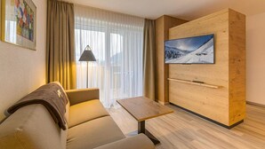 A modern hotel room with a sofa, a small table, a TV mounted on a wooden panel, and a window with curtains. - Residenz Schiestl (Fuegenberg)